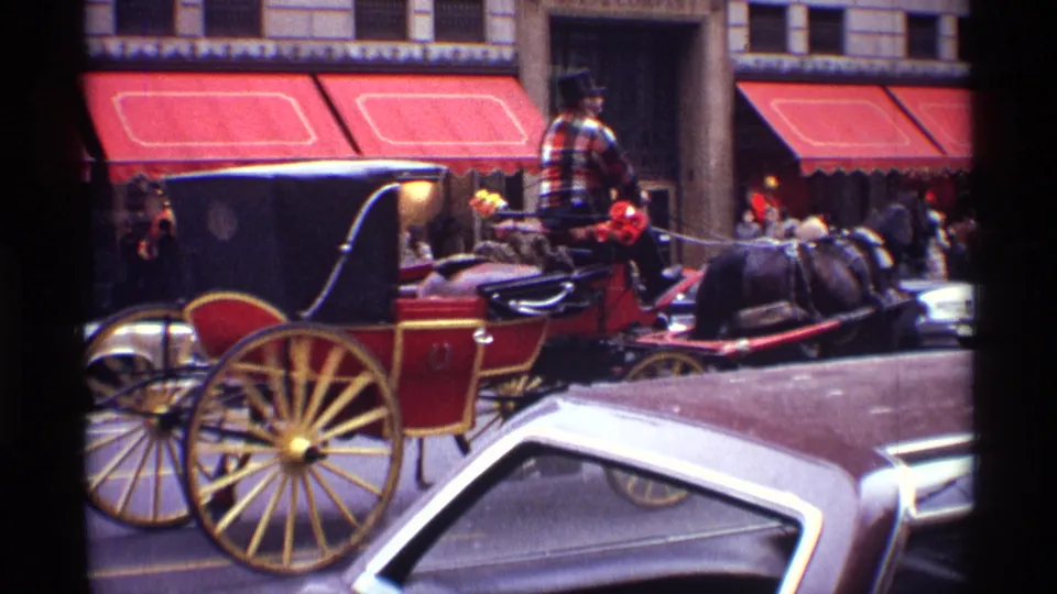 A red horse-drawn carriage with yellow wheels travels through a 1970 New York City street scene with cars and storefront awnings.