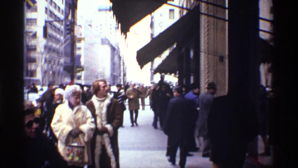Street-level 1970 New York City sidewalk view with pedestrians in coats walking between tall buildings and storefronts.