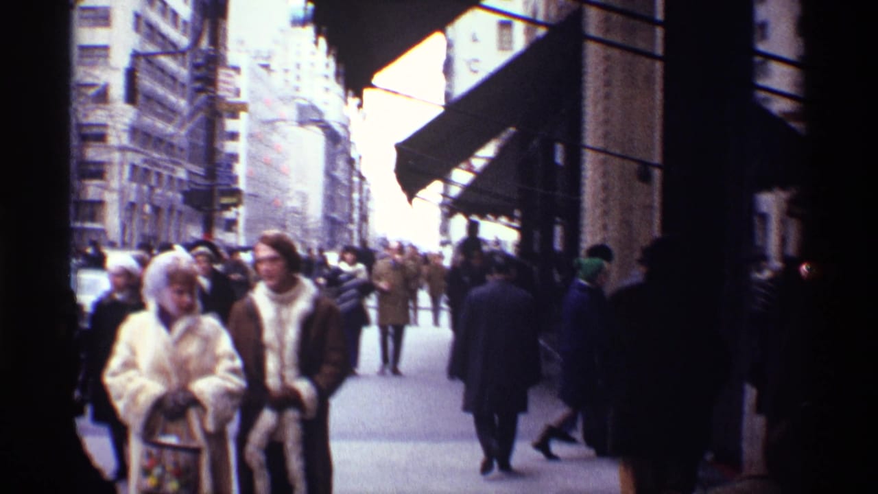 Pedestrians in winter coats walk along a busy city sidewalk beside tall buildings and awnings.