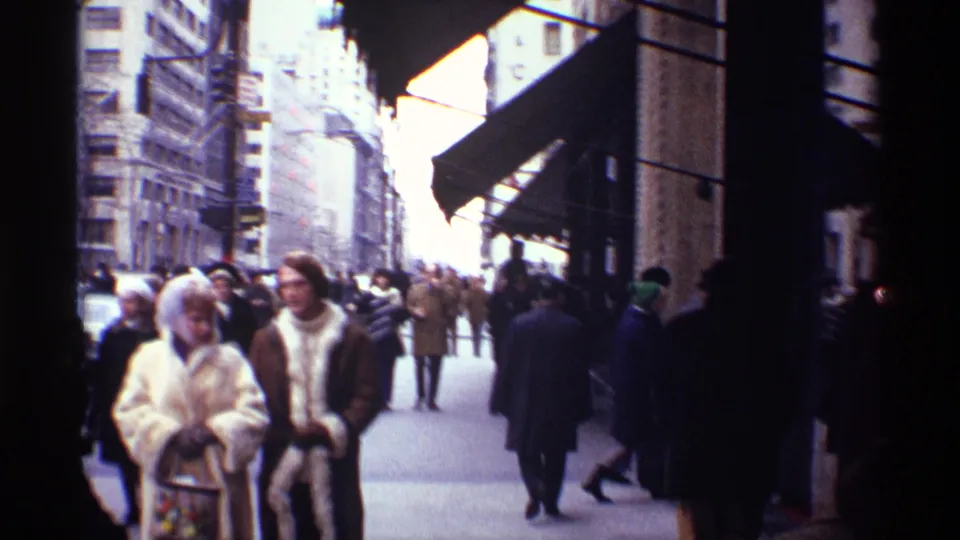 1970 New York City sidewalk scene with bundled pedestrians passing storefront awnings and tall building facades.