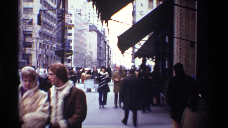 Two bundled pedestrians in 1970 New York City stand near the camera while a winter sidewalk crowd moves behind them.