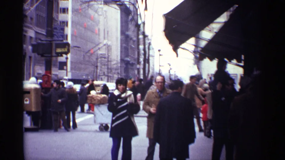 Winter sidewalk view in 1970 New York City with a striped-coat pedestrian, bundled walkers, storefront awnings, and street fixtures.
