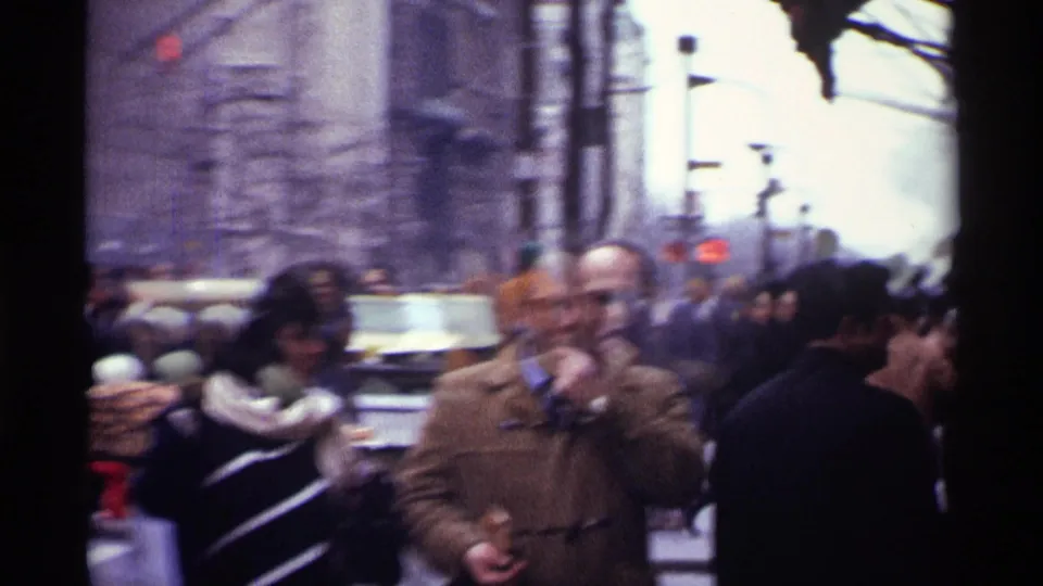 Close street-level 1970 New York City frame with a man in a brown coat, passing pedestrians, cars, and soft handheld motion blur.