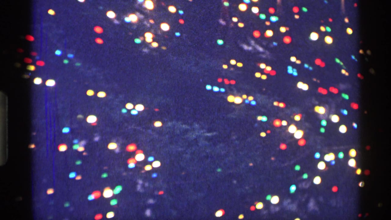 1970 New York City 8mm frame with soft red, green, blue, and white holiday lights blurred against dark branches.