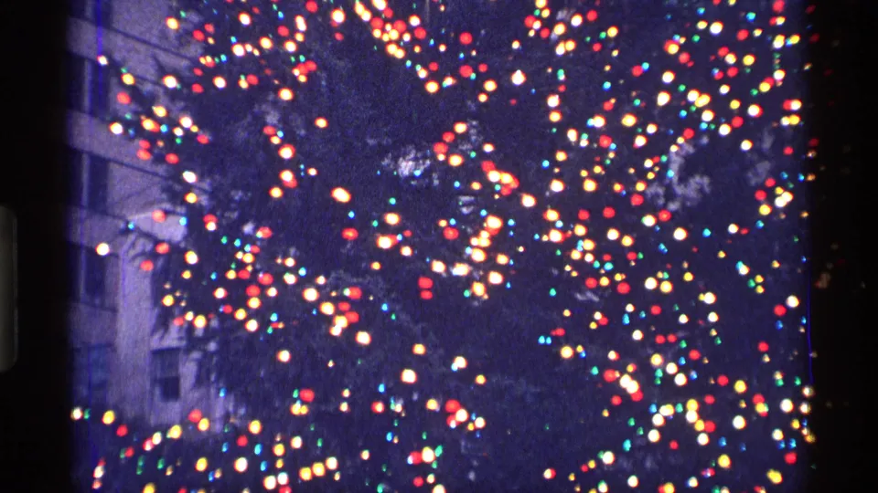 A large decorated Christmas tree glows with dense colored lights beside a tall building in 1970 New York City footage.