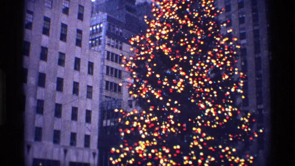 A tall city Christmas tree covered in multicolored lights stands beside high-rise buildings in 1970 New York City.
