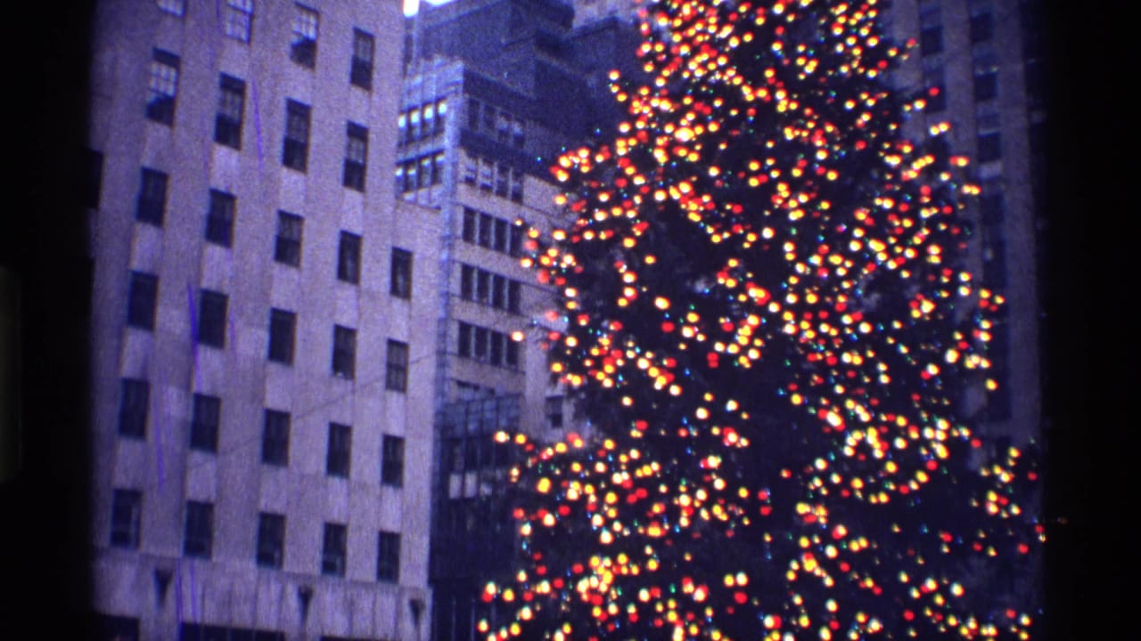 Lit Christmas tree beside tall city buildings at dusk.
