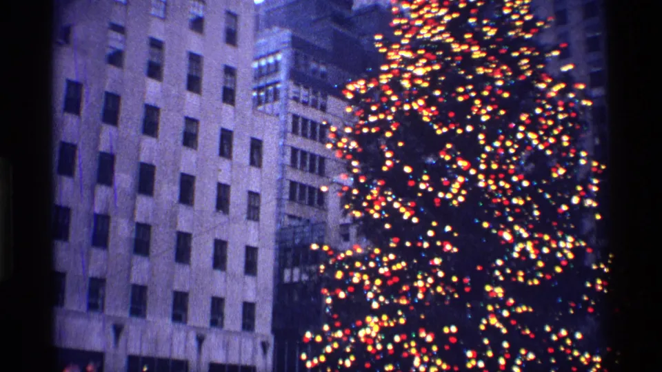 A close 8mm view of the Rockefeller Center Christmas tree glowing against tall New York City buildings in 1970.