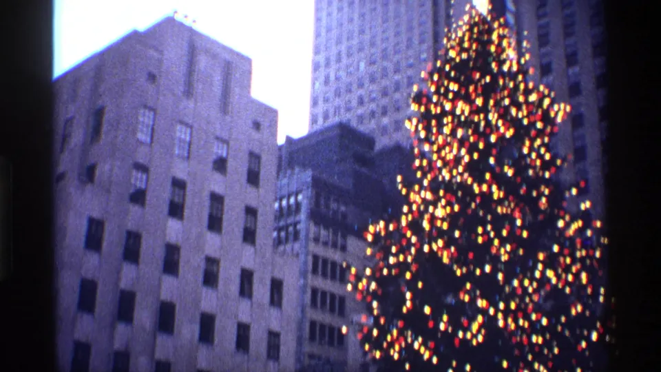 Low-angle 1970 New York City frame with the illuminated Rockefeller Center Christmas tree rising beside midtown towers.