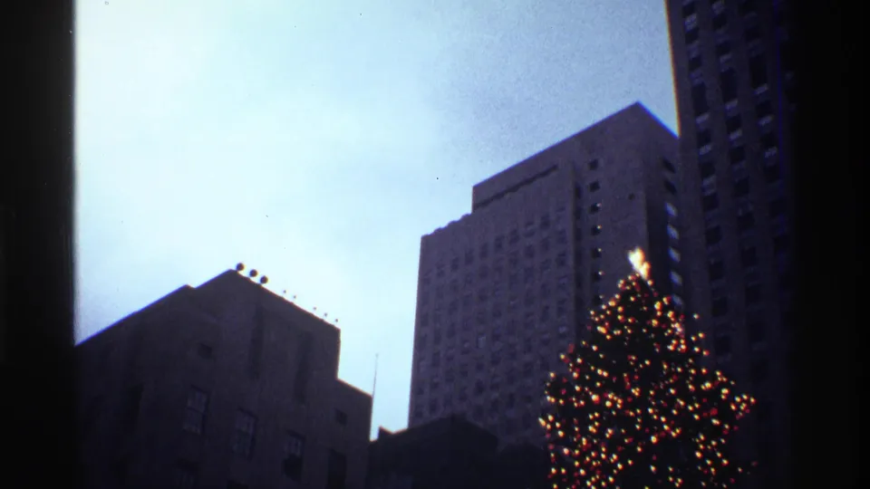 A wider low-angle 8mm frame shows the Rockefeller Center Christmas tree lights below dark New York City building silhouettes.