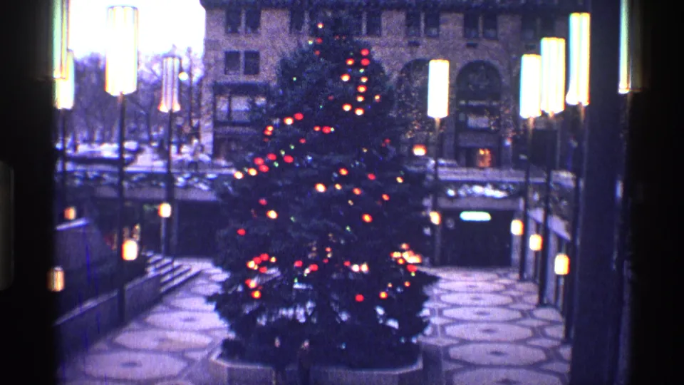 A large decorated Christmas tree stands in a New York City plaza, framed by tall lamps, patterned paving, and nearby buildings.