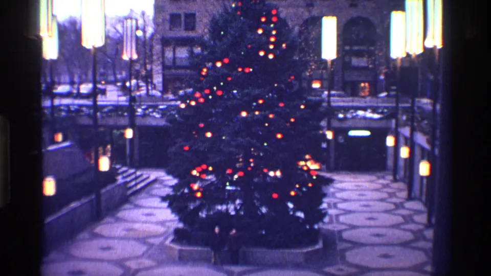 The illuminated Christmas tree fills the center of the frame, with plaza lamps, steps, paving patterns, and building facades around it.