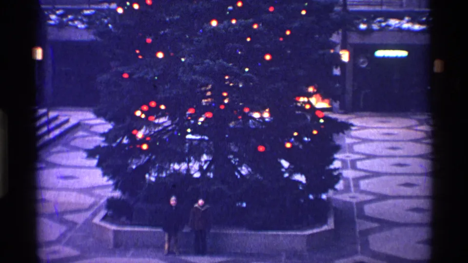 Two distant people stand at the base of the large decorated Christmas tree in the New York City plaza, with lights and paving visible.