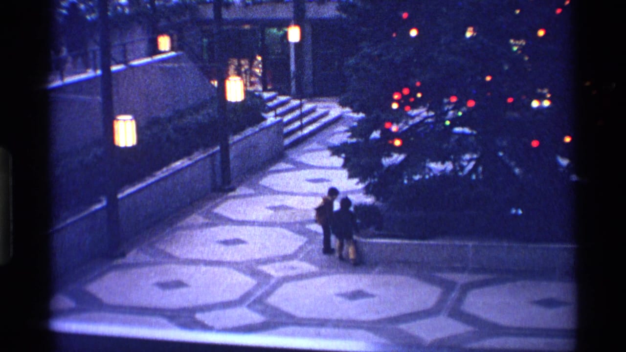 Two small figures stand on a patterned plaza near lantern lights and a lit tree at night.