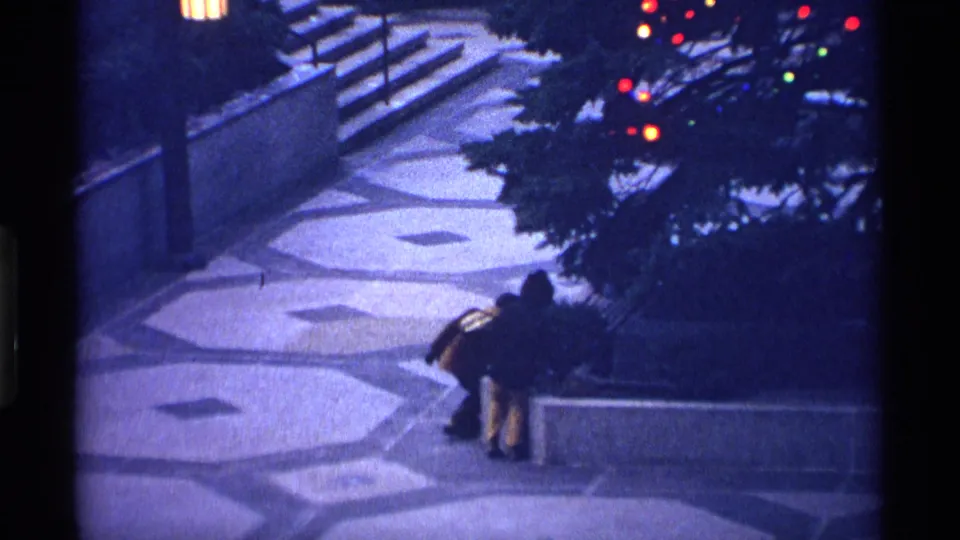 1970 New York City nighttime frame with two bundled children beside a low wall, steps, and a shadowed planting area.