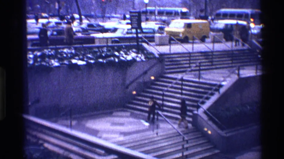 1970 New York City home-movie frame showing two children on concrete stairs below traffic, buses, parked cars, and bare trees.