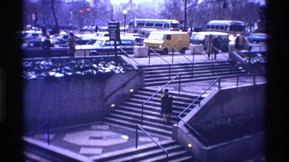 1970 New York City street view with a pedestrian on concrete steps, metal railings, buses, cars, and bare trees in the background.