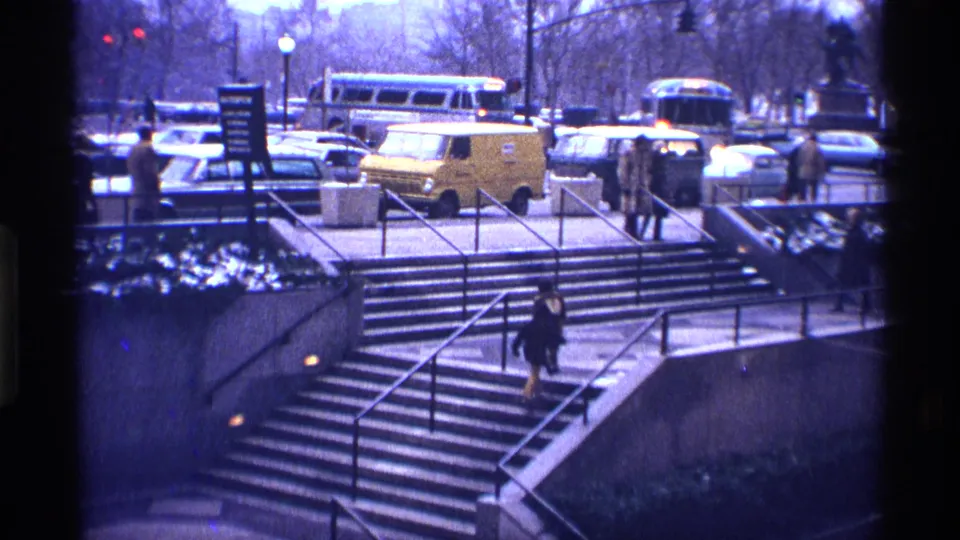 1970 New York City home-movie frame with a person on broad concrete stairs and period traffic visible above the plaza edge.
