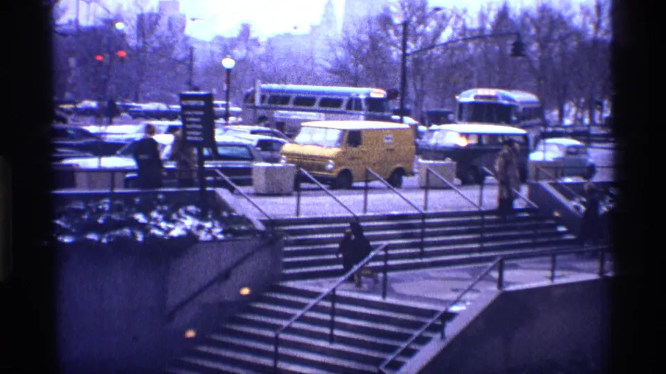 1970 New York City street scene viewed across concrete stairs, with pedestrians, buses, a yellow van, parked cars, and winter trees.