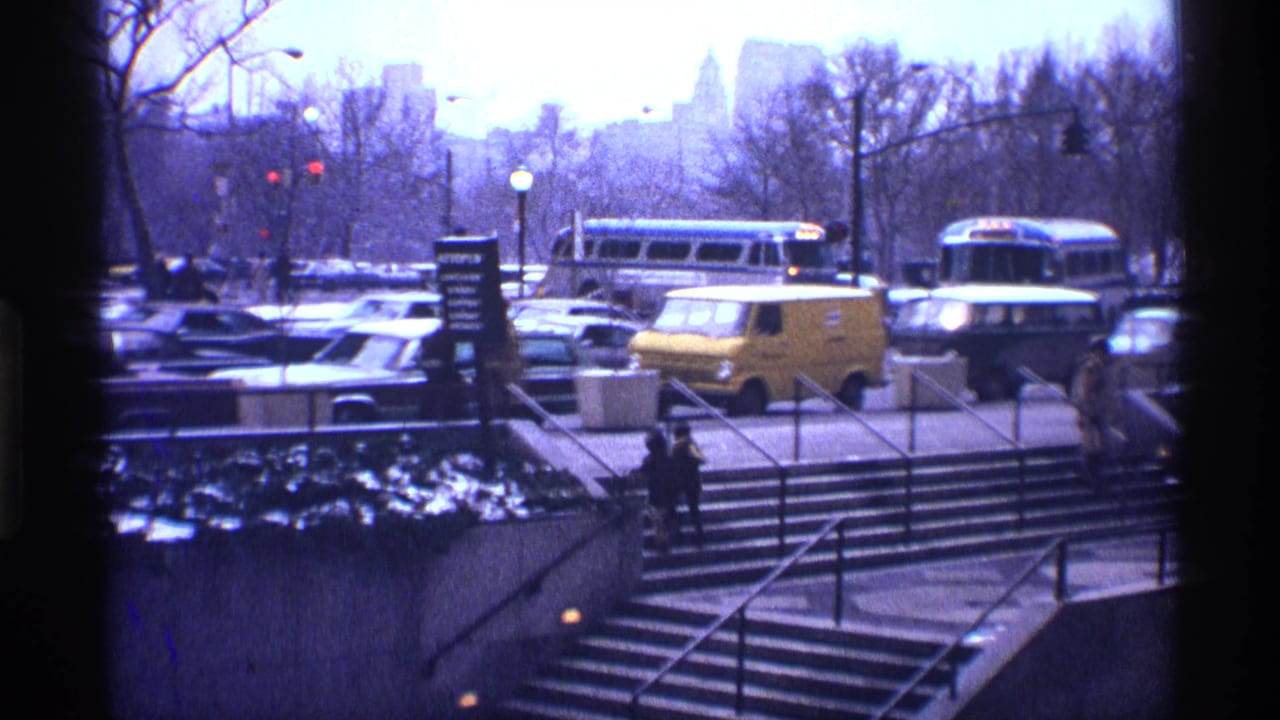 Busy street with cars, buses, a yellow van, subway steps, bare trees, and snow patches.