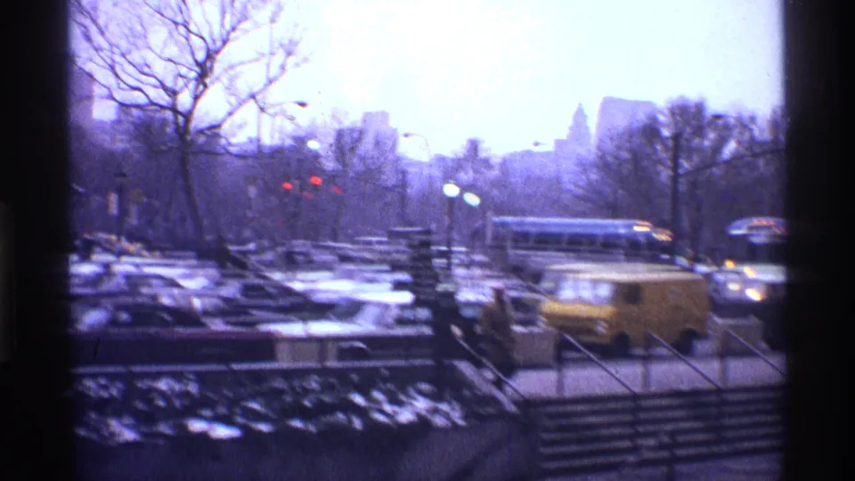 1970 New York City traffic scene with a bus, yellow van, cars, subway-area stair railings, bare winter trees, and hazy buildings beyond.