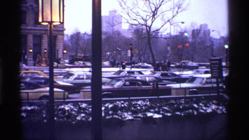 1970 New York City street view with rows of cars, tall lamps, bare trees, snow along the foreground, and pedestrians partly visible near the curb.