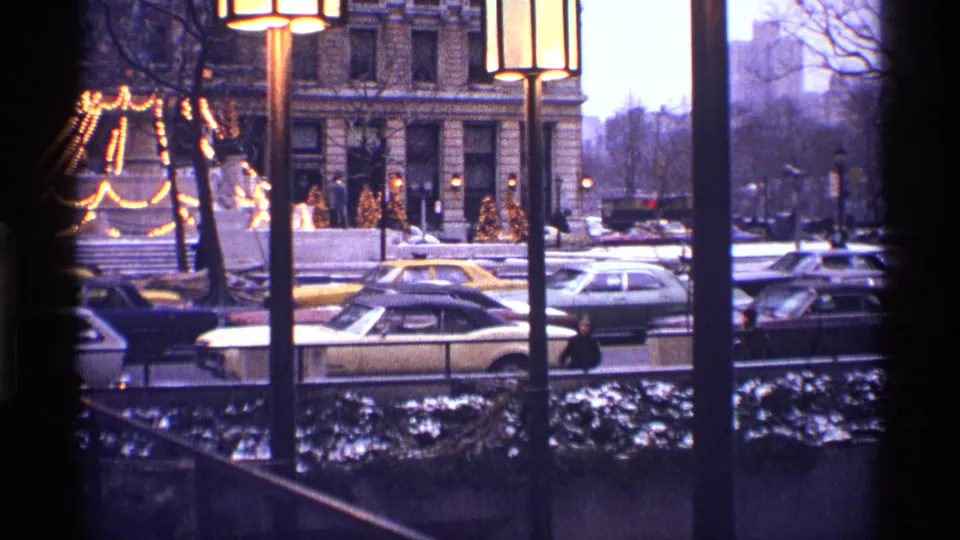 1970 New York City street traffic seen past tall lamps, an ornate building facade, bare trees, snow patches, pedestrians, and lighted decorations.