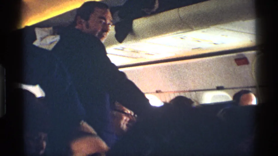 A man in a dark suit stands or leans in the aisle of a 1970 New York City railroad car while passengers sit nearby.