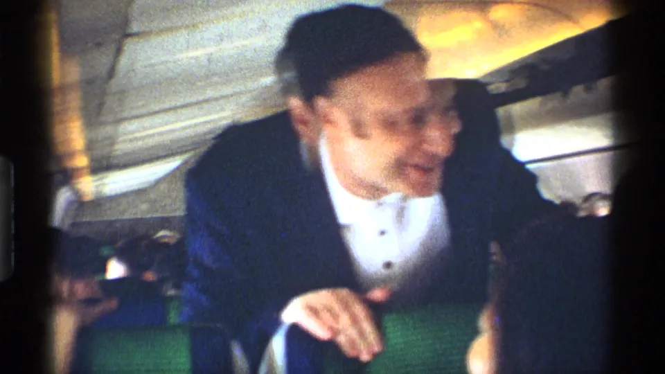 A suited man smiles toward the camera inside the 1970 New York City railroad car, with green seating and passengers around him.