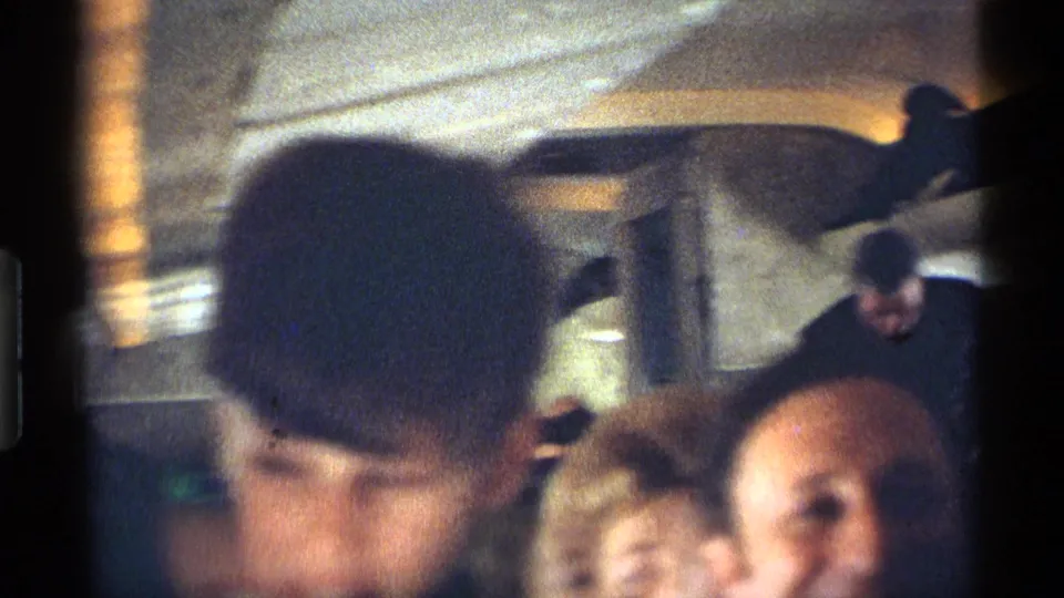The home-movie frame shows blurred passengers in a 1970 New York City railroad car, with a dark-haired person close to the camera.