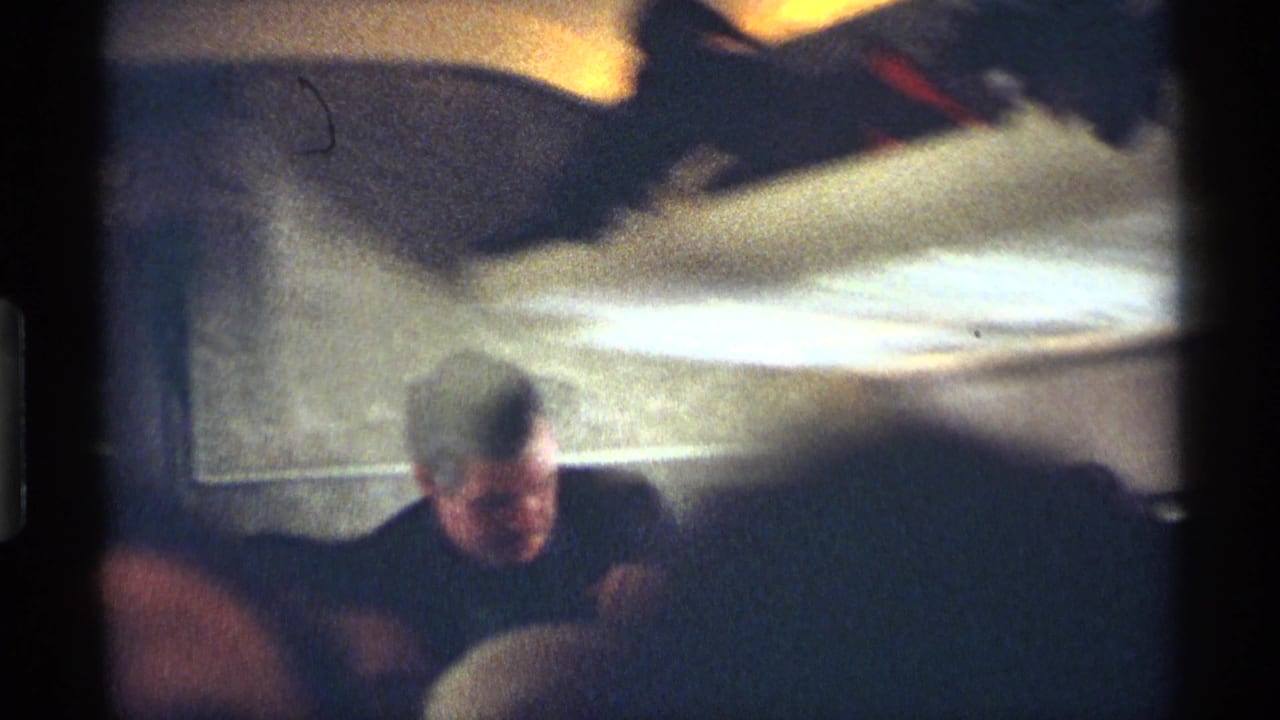 Seated airplane passengers beneath warm cabin lighting, with an adult man visible near the front of the frame.
