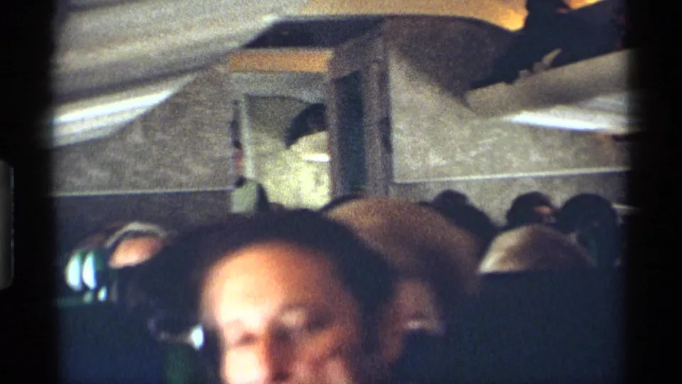 Handheld 8mm frame from inside a 1970 airplane cabin, showing green seat backs, seated passengers, overhead storage areas, and a soft close foreground face.