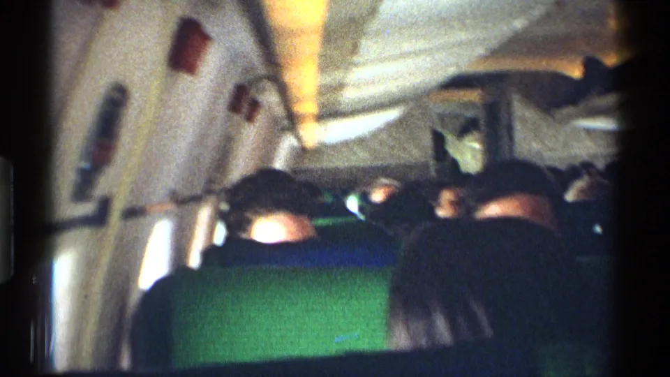 1970 8mm passenger-view frame looking along a commercial aircraft cabin, with green seat backs, window light, overhead panels, and seated travelers in partial silhouette.