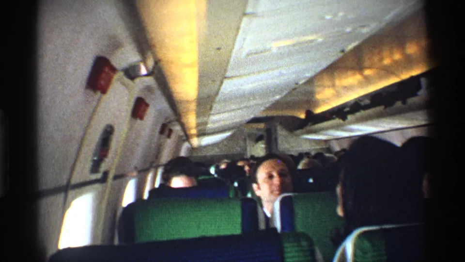 1970 airplane cabin footage with passengers in patterned seats, overhead bins above, and a man facing the handheld camera.