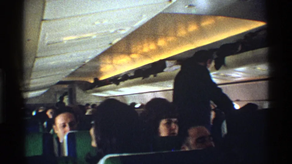 Home-movie frame inside a commercial airplane, showing passengers seated closely while one person stands below the overhead storage.