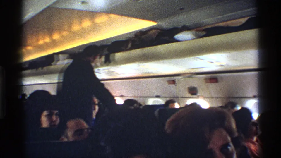 1970 cabin view from a passenger perspective, with a standing traveler near overhead bins and seated people visible below the window line.