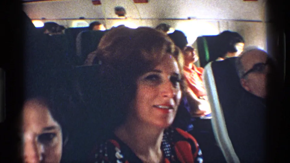 1970 8mm frame of a woman smiling toward the camera from an airplane seat, with rows of passengers and cabin panels visible.