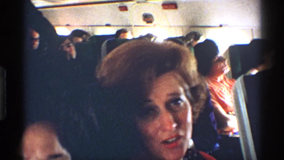 1970 8mm airplane-cabin frame with a closer view of a seated woman, other passengers, seatbacks, and overhead panels behind her.