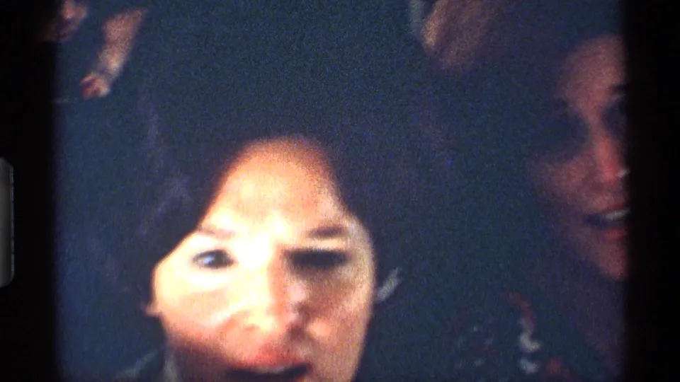 Soft 1970 8mm close-up inside the airplane cabin, with a woman's face near the lens and another passenger partly visible at right.
