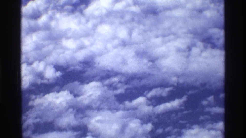 1970 8mm home-movie view of puffy clouds and blue sky through a dark airplane-window edge, with visible film grain.