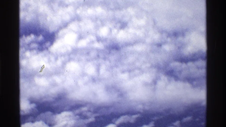 Cloudscape from an airplane-window perspective in 1970 8mm footage, showing soft focus, blue-white color, and small film marks.