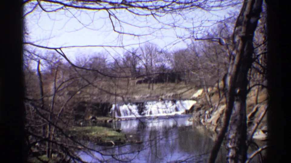 A wooded New York City winter scene shows white water over a low dam, viewed through dark tree trunks on 8mm film.