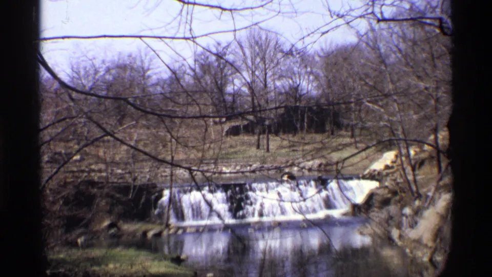 The camera moves closer to the small waterfall, with bare branches crossing a 1971 New York City winter waterway view.