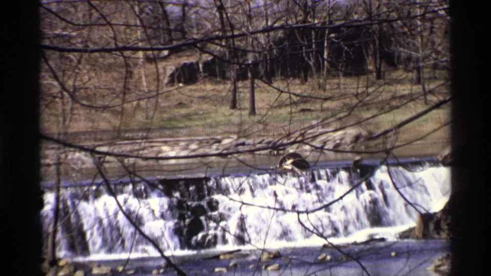 Close 8mm view of water spilling over a low dam in New York City, framed by winter branches and wooded banks.