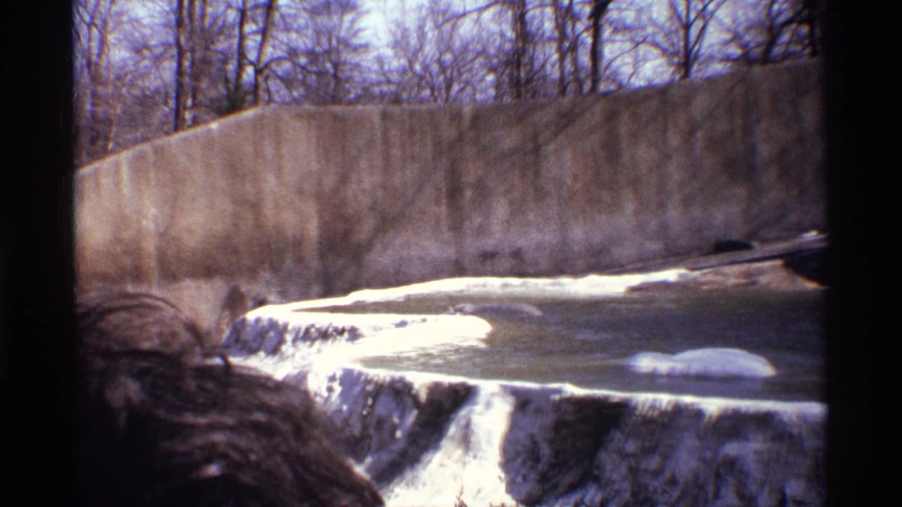 Polar bear lying low in a concrete zoo pool beside white ledges and a high wall.