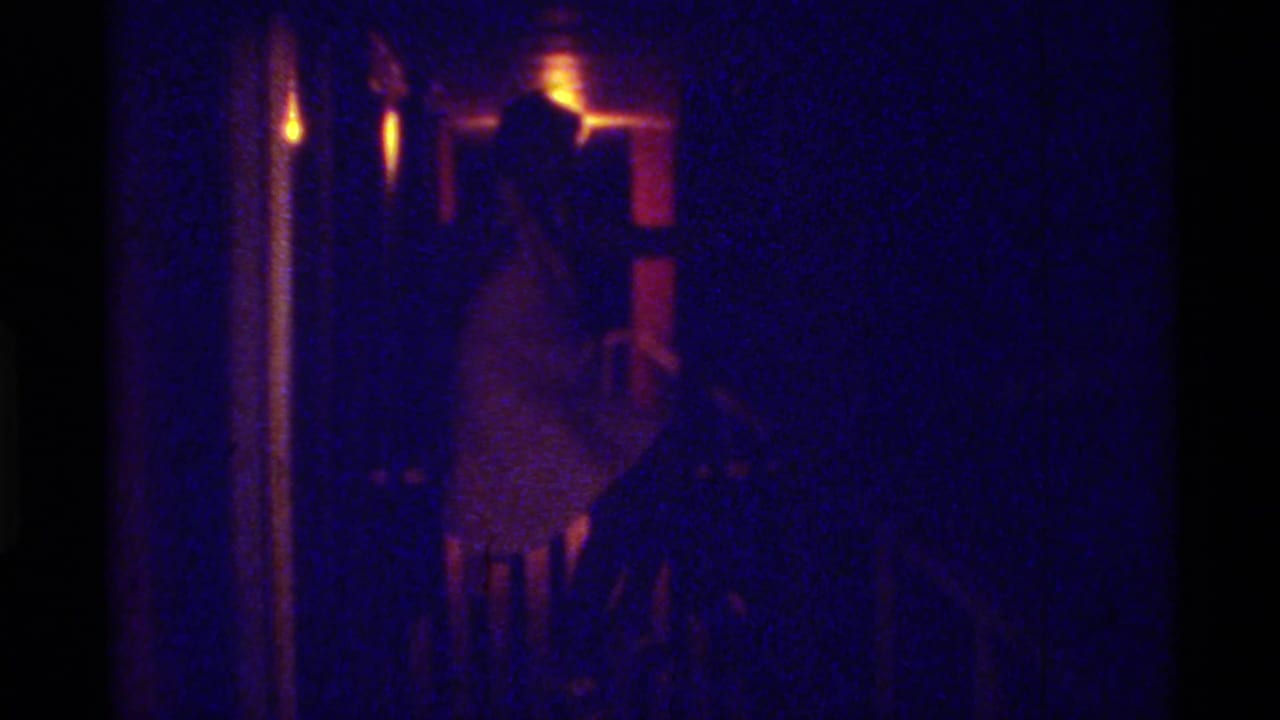 1973 Florida 8mm frame showing an adult woman in a light dress seated in a dark hall or stair area near a warm exposed light.