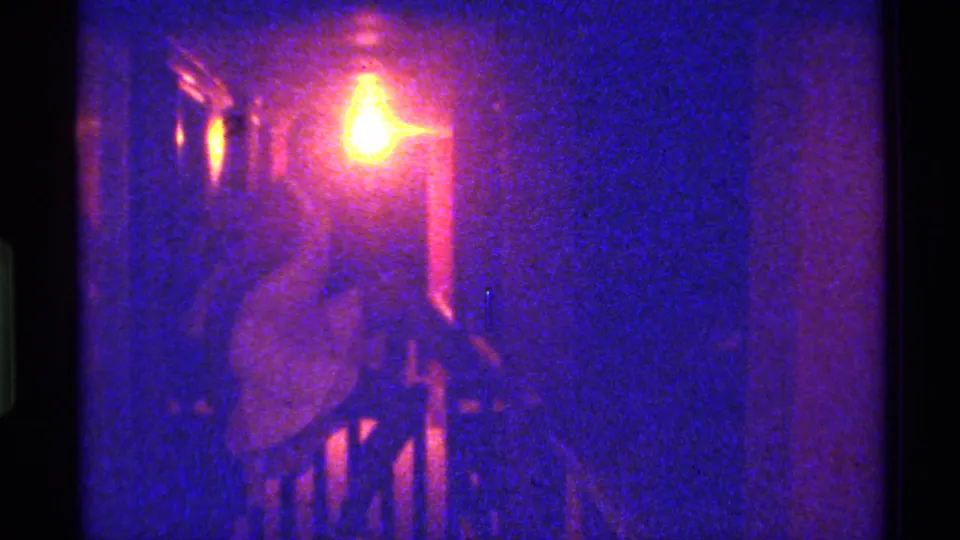 1973 Florida 8mm frame showing the woman still seated near a railing in a dim private interior with warm light and purple-blue color cast.
