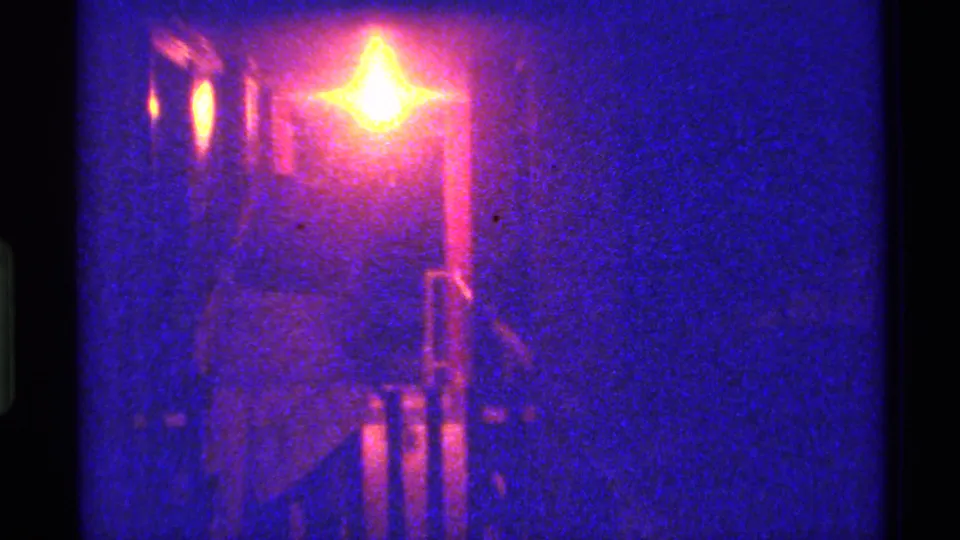Blue-toned 1973 Florida home-movie frame with a glowing lamp and indistinct railing or gate forms in darkness.