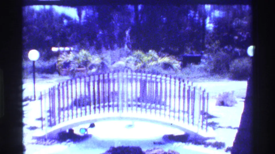 1973 Florida 8mm frame of a sunlit landscaped park with a curved wooden bridge, globe lights, shrubs, and trees.