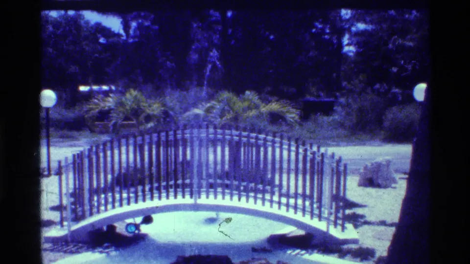 1973 Florida home-movie frame showing a curved park bridge, dark trees, globe lights, and landscaped shrubs in bright daylight.